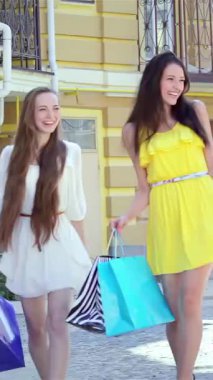 Coşkulu üç genç kadın, her biri şık kıyafetler ve renkli parlak alışveriş torbalarıyla gösterişli bir caddede geziniyor. Bulaşıcı gülüşleri ve ışıl ışıl gülümsemeleri arkadaşlığı kutlayan saf bir neşe ortamı yaratıyor.