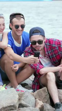 Üç canlı arkadaş, iki erkek ve bir kadın, güneşli kayalık kıyılarda bir araya gelerek selfie çekerken mutluluk saçıyorlar. Kahkahaları rüzgarda dans eder, suyun ışıl ışıl arka planına karşı anılar yaratır.