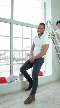 İki canlı genç müteahhit, biri kendine güvenen bir merdivene tünemiş ve diğeri büyük bir pencerenin yanında duruyor, boya kutularını tutuyor ve hevesle başparmaklarını kaldırıyor. Renkli kaskları ve günlük kıyafetleri parlak, havadar iç mekanlarına çarpıyor.