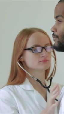 Odaklanmış, kızıl saçlı bir kadın doktor erkek hastalarının kalp atışlarını steteskop kullanarak nazikçe dinliyor ve şık, modern bir klinikte sıcak ve şefkatli bir ortam yaratıyor. Bu samimi an hayatî bağlantıyı vurguluyor.