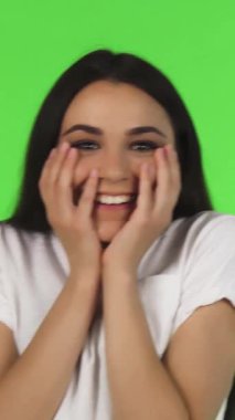 Çekici, genç, esmer bir kadın sevinçle ışıldıyor elleri neşe dolu bir sürpriz anında yüzünü çerçeveliyor. Canlı yeşil bir arkaplan oluşturan neşeli ifadesi, olumlu ve kutlayıcı bir şekilde izleyicileri katılmaya davet ediyor.