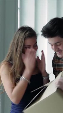 Genç bir çift, güneşli bir pencerenin yanında duruyor, bir kutu tutuyorlar ve yeni dairelerinde paketlenmemiş karton kutularla çevrili, parlak gülümsemelerle dışarı bakıyorlar. İfadeleri saf neşe ve heyecan yayar, hayata atılmanın heyecanını somutlaştırır.