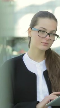 A poised and confident businesswoman, dressed in sleek formal attire, stands outdoors, intently reviewing her tablet while preparing to welcome new partners. Her thoughtful expression and professional demeanor, framed by a striking modern
