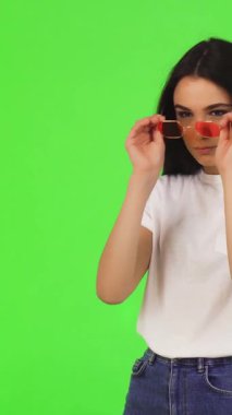 A striking young woman with long, flowing dark hair playfully adjusts her trendy pink sunglasses, set against a vivid green backdrop that radiates energy. Her casual white tee and relaxed demeanor create a chic, inviting atmosphere, encouraging