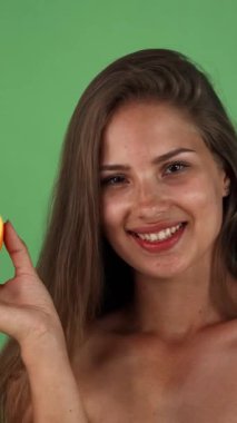 A radiant young woman with long, flowing hair beams with joy as she playfully holds a vibrant half orange, symbolizing health and vitality. Set against a lively green backdrop, this image captures the essence of wellness and the delightful joy of