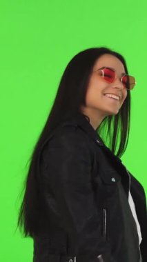 A radiant young woman exudes happiness as she playfully poses with her stylish shopping bags on a vivid green chromakey background. Clad in a chic leather jacket and trendy sunglasses, she captures the essence of youthful exuberance and modern