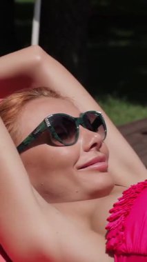 A stunning Caucasian woman with vibrant red hair luxuriates under the golden sun, reclining elegantly on a deck-chair beside the sparkling pool. With stylish sunglasses resting on her nose, she playfully glides her hand along her toned body