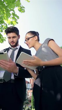 A confident businessman and a successful businesswoman engage with a tablet, their faces illuminated by the sun, surrounded by vibrant greenery and sleek modern architecture. Their joyful expressions radiate the excitement of achievement, embodying a