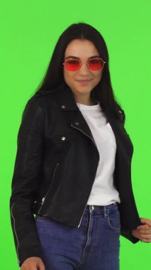 A fashionable woman with long dark hair and trendy sunglasses beams with joy as she holds a chic shopping bag towards the camera, set against a vibrant green chromakey background. This dynamic portrait encapsulates the excitement of retail therapy