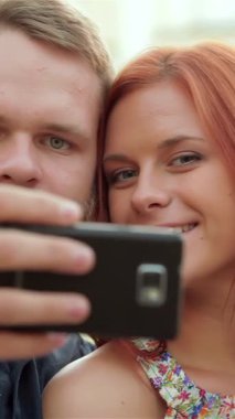 A cheerful young couple, the woman with radiant red hair and the man sporting a trendy beard, are joyfully taking a selfie with their smartphone while seated on a sunlit city bench. Their bright smiles shine against a colorful urban backdrop