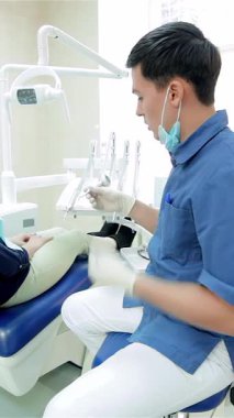 A dedicated young dentist, clad in a crisp blue uniform, attentively interacts with a patient seated in a sleek dental chair, radiating warmth and professionalism. The bright, contemporary clinic is filled with gleaming dental tools, creating an