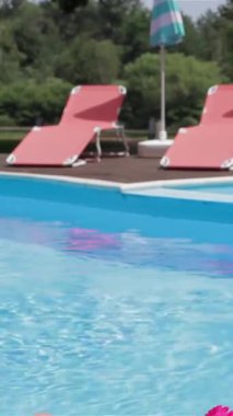 A stunning Caucasian woman with flowing ginger hair reclines gracefully on a sunbed, soaking in the warm rays while wearing a vibrant pink bikini. The scene radiates summer bliss, with the sparkling turquoise water reflecting the azure sky, creating