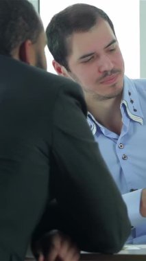 Two focused businessmen engage in a lively discussion, analyzing reports with intent expressions at a modern negotiation table. Dressed in sharp formalwear, they radiate professionalism and collaboration, surrounded by a bright office space that