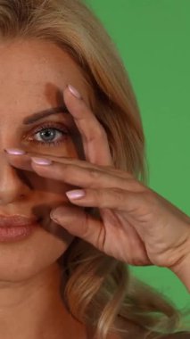 This striking half-face portrait captures the radiant beauty of a mature woman, gently caressing her cheek with a perfectly manicured hand against a vibrant green chromakey backdrop. The image exudes confidence and sophistication, celebrating the