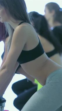 An athletic girl in a sleek black sports bra and form-fitting gray leggings pedals with fierce determination on a stationary bike, surrounded by her equally committed friends at the gym. The bright, airy environment radiates energy, highlighting