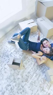 In a bright, airy apartment, a couple joyfully lies on the floor in a loving embrace, surrounded by scattered unpacked boxes. Their radiant smiles and cozy attire reflect the thrill of embarking on a new journey together in their fresh, inviting