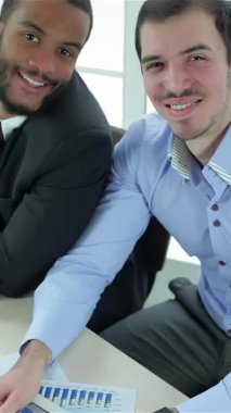 In a sunlit modern office, two successful businessmen, one in a sharp blue shirt and the other in a tailored suit, beam with pride while giving a thumbs up. Their vibrant smiles and confident demeanor exude positivity and teamwork, capturing a