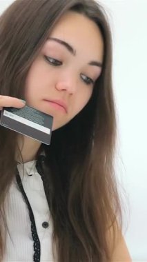 A contemplative young woman with flowing brown hair examines her credit card while intently browsing her laptop, fully engaged in the thrill of online shopping. The bright, minimalist backdrop accentuates her focused expression, capturing the