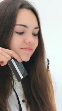 A cheerful young woman with long, flowing hair beams as she prepares to make an online purchase, holding her sleek credit card close to her face. The bright, minimalist background radiates a sense of excitement and modernity, perfectly capturing the