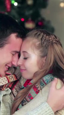 In a heartwarming Christmas scene, a loving couple, adorned in snug sweaters and vibrant scarves, gently touch foreheads, radiating warmth and affection. Surrounded by twinkling lights and a beautifully adorned tree, this intimate moment captures the