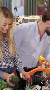A cheerful young couple is joyfully picking out vibrant vegetables at the supermarket, surrounded by a kaleidoscope of fresh produce. The handsome Caucasian man, sporting a stylish beard, playfully hands a ripe avocado to his blonde girlfriend, whose