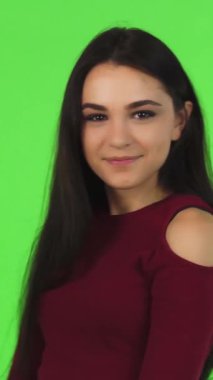 A vibrant young woman with long, flowing brunette hair joyfully points at the camera, her bright smile lighting up the scene against a lively green backdrop. Her infectious energy and confident demeanor invite viewers to share in her excitement