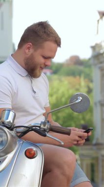 A cheerful young man with a stylish beard sits casually on his sleek scooter, deeply engaged in a lively conversation on his smartphone. The vibrant urban landscape, bathed in warm sunlight, amplifies the sense of freedom and adventure, making this