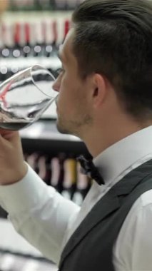 A dapper young man in a sleek waistcoat and bow tie gracefully sips from a crystal glass, surrounded by a dazzling array of wine bottles in a chic liquor store. His confident posture and discerning gaze reflect the artistry of winemaking, inviting