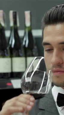 A sophisticated male sommelier in a sharp tuxedo leans over a polished wooden table, deeply inhaling the rich aroma of a luxurious red wine. Behind him, an elegant display of fine wine bottles creates an upscale ambiance, perfect for this refined