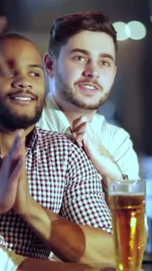 Two young businessmen, one with a beard and the other with a stylish haircut, share a moment of excitement as they cheer for their favorite football team, raising their glasses of golden beer. The bar buzzes with energy, illuminated by warm lights