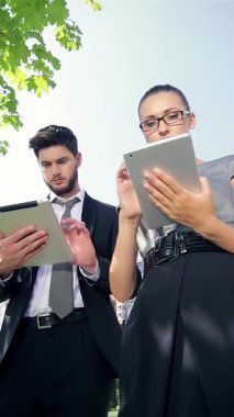 Odaklanmış bir işadamı ve kararlı bir iş kadını, modern mimari ve canlı yeşillik çerçeveli canlı bir şehir parkında tabletleri üzerinde işbirliği yapıyorlar. Keskin giysileri ve nişanlı ifadeleri profesyonelliği ve yeniliği yansıtıyor.