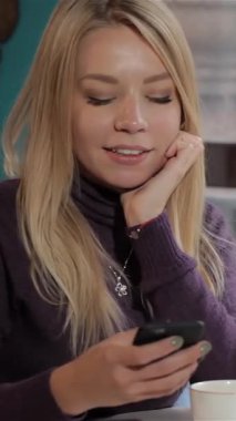 Güzel bir kafede oturmuş mutluluk saçan, akıcı sarı saçlı, mutlu bir beyaz kadın akıllı telefonuna sarılmış durumda. Çekici atmosfer, onun sıcak gülüşü ve dumanı tüten bir fincan kahveyle mutlu bir an yaratıyor.