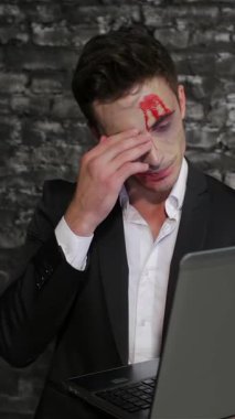 Pürüzsüz siyah bir takım elbise giymiş büyüleyici bir erkek vampir laptopunu yoğun bir şekilde inceliyor, kan kırmızısı alnındaki sembolü, desenli siyah tuğla bir duvara karşı kaygı verici bir şekilde parlıyor. Bu çarpıcı görüntü modern teknolojiyle klasik korkuyu birleştiriyor.