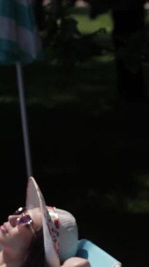 İki muhteşem beyaz kadın, biri esmer, diğeri kızıl saçlı, parlak bir havuzun yanında canlı turkuaz şezlonglarda güneşleniyorlar. Şık bikiniler ve geniş kenarlı şapkalarla süslenmiş bir rahatlama ve neşe havası yayarlar.