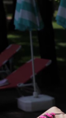 İki muhteşem beyaz kadın, biri esmer ve diğeri ateşli kızıl saçlı, ışıl ışıl bir havuzun yanındaki iskemlede güneşleniyorlar. Modaya uygun bikiniler ve şık plaj şapkaları giyip rahatlama ve neşe saçarak sıcacık havayı emiyorlar.