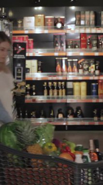 Mutlu bir çift, canlı bir hipermarket koridorunda raflardan renkli içecekler seçiyor. Genç beyaz erkek ve kadın, neşe saçan, taze ürünlerle dolu bir alışveriş arabasını tutuyor, heyecanını mükemmel bir şekilde yakalıyor.