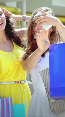 Üç modaya uygun kız, her biri gösterişli renkli alışveriş torbaları, şık bir alışveriş merkezinin dışında canlı bir selfie paylaşıyorlar. Işıldayan gülümsemeleri ve neşeli jestleri, dostluk, moda ve eğlenceyle dolu bir günün heyecanını yayıyor.