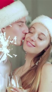 Neşeli Noel Baba şapkaları takan hoş bir çift, maytap tutarken tatlı bir öpücük paylaşıyor, yüzlerini neşeyle aydınlatıyor. Bayram ışıklarının büyüleyici parıltısı ve güzel süslenmiş Noel ağacı sıcak, samimi bir Noel ağacı yaratır.