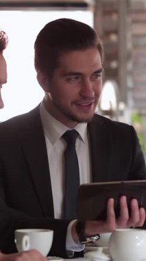 Üç iyi giyimli adam şık bir kafede lezzetli bir öğle yemeğinde stratejileri tartışırken gülümsemeleri güven ve dostluk saçıyor. Sıcak kahve ve parlak bir tabletle dolu davetkar atmosfer dinamiklerini vurguluyor.