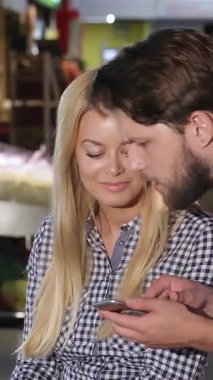 Alışverişe dalmış şık genç bir çift alışverişlerinin fiyatını canlı bir süpermarkette hesaplıyor. Sakallı adam akıllı telefonuna dikkatle odaklanırken sarışın kadın neşeyle ışıldıyor.