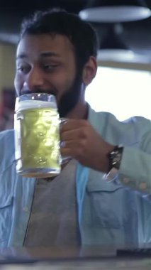 Neşeli bir erkek futbol fanatiği soğuk bira bardağını hareketli bir barda kaldırıyor, heyecan ve dostluk saçıyor. Neşeli atmosfer kahkahalarla uğulduyor ve maçın heyecanıyla, arkadaşlar soğuk içeceklerle futbol için paylaştıkları tutkuyu kutluyorlar.