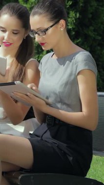 In a lively outdoor setting, two fashionable young businesswomen are deeply engaged in their calls, radiating a vibrant energy of collaboration. Their chic outfits and confident expressions reflect a modern corporate culture, while a third colleague