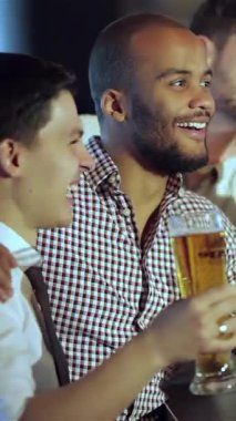 Two excited friends, a Black man and a Caucasian man, share a hearty laugh while raising their frosty beer glasses in a lively bar, immersed in the thrilling football match on screen. The vibrant atmosphere buzzes with camaraderie and joy, as they