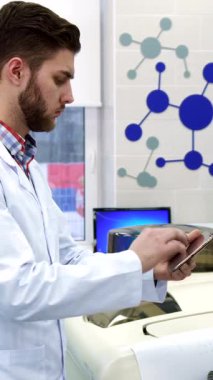 A dedicated young Caucasian scientist in a crisp white lab coat meticulously transfers data from a computer to a sleek tablet, surrounded by cutting-edge laboratory equipment. His thoughtful expression and the vibrant blue molecular graphics on the