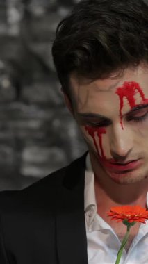 A male vampire in a sleek black suit, with blood-red streaks on his pale face, hauntingly tears petals from a vibrant orange flower against a shadowy brick wall. This eerie scene encapsulates the spirit of Halloween, merging gothic horror with an air