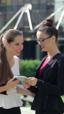 A poised young businesswoman stands outside a sleek modern office, intently engaging with her tablet before breaking into a radiant smile for the camera. Surrounded by fellow professionals in stylish attire, her confident demeanor and vibrant energy