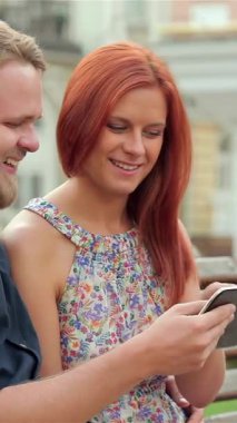 A radiant couple sits closely on a rustic wooden bench, captivated by their tablet as laughter and smiles light up their faces in a bustling urban park. Bathed in warm sunlight, their joyful connection radiates the essence of summer adventures