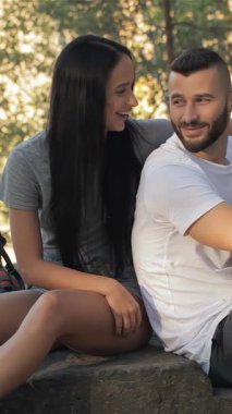 A vibrant couple enjoys a delightful moment on a sun-kissed rock in a lush forest, engaging in animated conversation. The brunette woman beams with joy, resting her hand on her boyfriends shoulder as he flashes a thumbs-up, embodying the spirit of