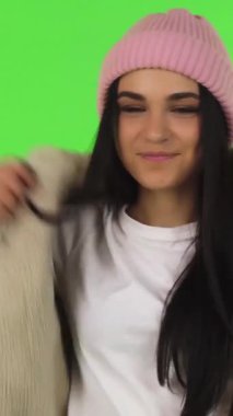In a vibrant studio with a striking green background, a cheerful young woman playfully puts on a cozy jacket, exuding warmth and positivity. Her charming smile and trendy pink beanie create an inviting atmosphere, perfectly capturing the essence of