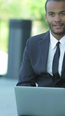 A charismatic businessman in a tailored suit sits outdoors, engaging with his laptop, radiating professionalism and warmth. The lush greenery surrounding him enhances the vibrant atmosphere of success and collaboration, inviting new opportunities and